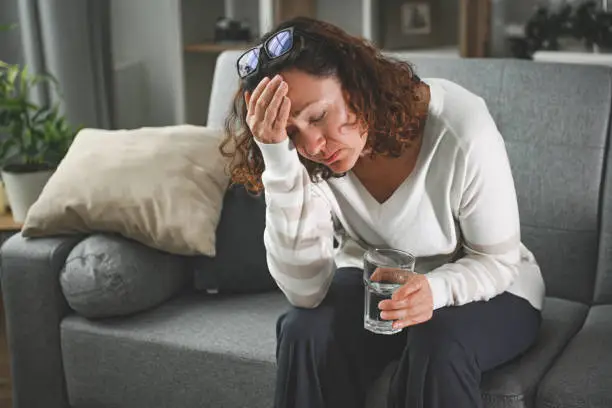 Woman experiencing heat stroke symptoms — sitting on couch, holding head and water glass, showing heat-related illness warning signs USA 2026