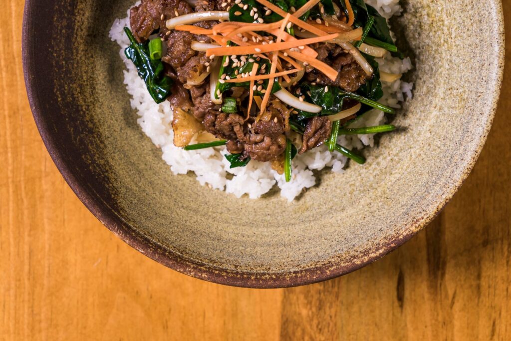 Overhead shot of a high-protein beef power bowl with white rice, sauteed beef strips, carrots, greens, and sesame seeds in a ceramic bowl