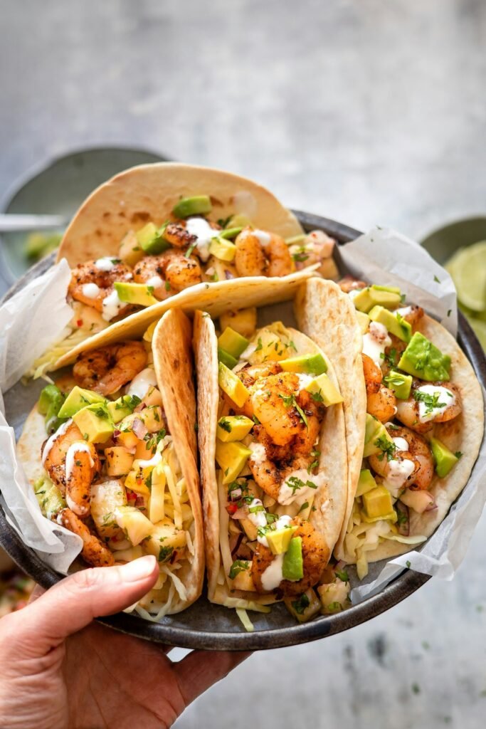 Hand holding a tray of chili-lime shrimp tacos topped with avocado and lime crema